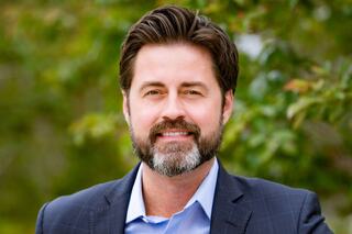 A smiling man with a beard and styled hair, dressed in a blue shirt and dark blazer, stands against a backdrop of greenery.