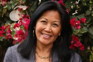 A woman with long, dark hair smiles amid vibrant pink flowers, wearing a gray blazer and elegant jewelry.