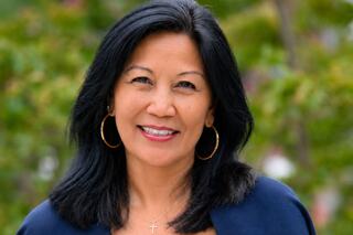 A woman with long black hair, wearing hoop earrings, smiles warmly against a blurred green background. She exudes confidence and friendliness.