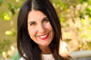 A smiling woman with long, dark hair and bright red lipstick poses in a sunlit outdoor setting with greenery in the background.