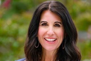 A woman with long, dark hair smiles warmly, wearing hoop earrings, against a blurred natural background. She exudes confidence and friendliness.
