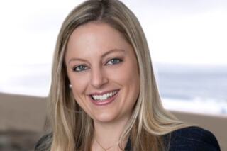 A woman with long blonde hair smiles warmly, wearing a dark blazer. The ocean and sandy beach are softly blurred in the background.