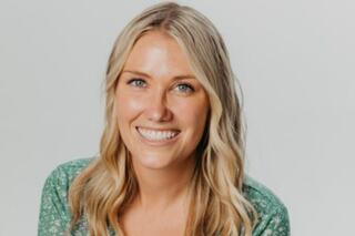 A smiling woman with long, wavy blonde hair wears a green floral top, radiating warmth and positivity against a neutral background.