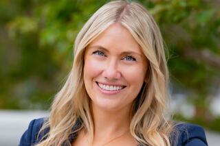 A smiling woman with long, blonde hair stands outdoors, radiating warmth and confidence against a blurred green background.