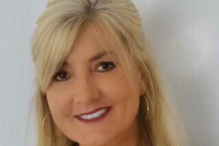 A smiling woman with long blonde hair and hoop earrings stands against a light background, exuding warmth and approachability.