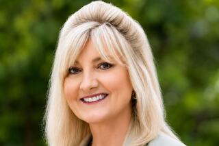 A smiling woman with long blonde hair styled elegantly, wearing a light-colored jacket against a blurred green background.