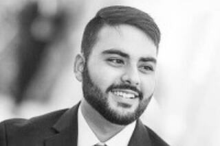 A smiling man in a suit, exuding confidence and warmth, with a neatly groomed beard and styled hair, set against a softly blurred background.
