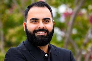 A smiling man with a thick beard wears a black suit, exuding confidence amidst a blurred green and pink background.