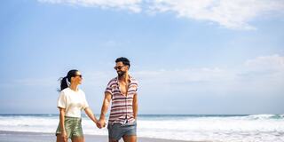 A couple strolls hand in hand along a sunny beach, smiling and enjoying the ocean breeze under a bright blue sky.