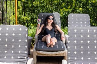 A woman in a black swimsuit relaxes on a lounge chair, holding a cocktail, surrounded by greenery and stylish chairs.