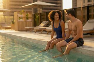 A smiling couple relaxes by a pool at sunset, their feet in the water, enjoying a warm evening in a stylish urban setting.