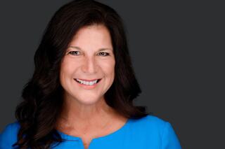 A smiling woman with long, dark hair wears a bright blue top against a dark background, exuding positivity and warmth.