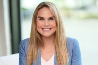A smiling woman with long, straight hair, wearing a light top and blue blazer, poses confidently in a bright, modern setting.