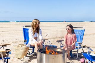 Two people enjoy a beach day, sitting by a fire pit. One toasts marshmallows, while the other looks on, both relaxed in beach chairs.