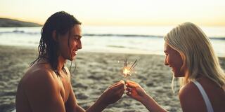 Two people share a sparkler at sunset on the beach, smiling and enjoying the moment together in a warm, romantic setting.