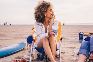A smiling woman with curly hair relaxes on a beach in a chair, holding a drink, while friends enjoy their time nearby.
