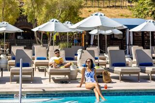 A couple strolls hand-in-hand through lush vineyards, each holding a glass of wine, enjoying a sunny day together.
