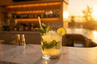 A refreshing cocktail with mint and lime sits on a marble counter, illuminated by warm sunset light, with a blurred bar in the background.