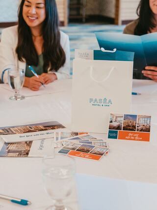A stylish meeting setup with a white bag from Paséa Hotel & Spa and promotional postcards, alongside individuals engaged in conversation.