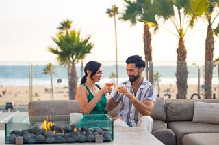 A couple toasts with drinks on a cozy outdoor sofa, surrounded by palm trees and a beach view, enjoying a warm, romantic evening.