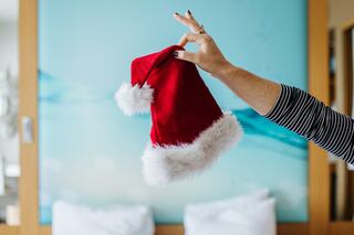 A hand holds up a festive red Santa hat with white fur trim against a soft blue background, evoking a cheerful holiday spirit.
