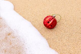A shiny red Christmas ornament lies on sandy beach, with gentle ocean waves lapping nearby, creating a unique holiday contrast.
