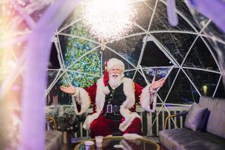 A joyful Santa Claus sits inside a festive igloo, surrounded by holiday decorations, warmly welcoming guests with open arms.