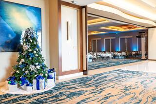A decorated Christmas tree with blue and silver ornaments stands beside a doorway leading to a stylishly arranged event space.