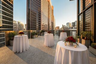 A stylish rooftop terrace features round tables with white tablecloths, floral centerpieces, and tall buildings under a clear sky.