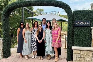 A group of eight individuals smiles together outside, dressed in various colorful outfits, standing under a decorative archway.