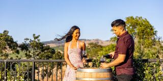 A couple enjoys wine on a sunny day at a scenic vineyard, with rolling hills and greenery in the background, creating a romantic atmosphere.