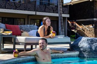 Two people relax by a pool, enjoying drinks and smiles under the sun, with a cozy lounge area and tropical ambiance surrounding them.