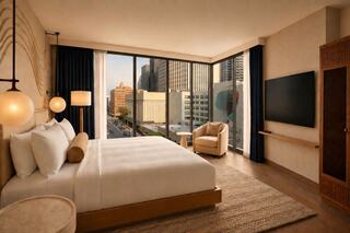 A modern hotel room features a large bed, stylish decor, and floor-to-ceiling windows with a cityscape view. Cozy and inviting atmosphere.