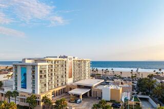 A modern hotel stands near the beach, surrounded by palm trees, with a clear blue sky and ocean in the background.