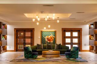 A stylish lobby features a green sofa, unique patterned chairs, wooden doors, shelves filled with decor, and modern lighting accents.