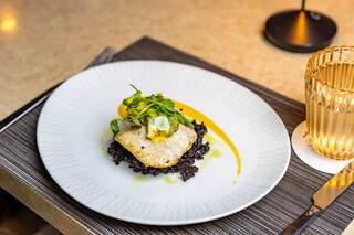 A beautifully plated dish features a piece of fish atop black rice, garnished with fresh greens and a drizzle of orange sauce.