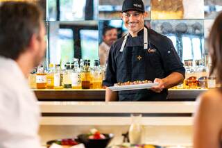 A smiling chef presents a dish to diners at a stylish restaurant, with shelves of liquor visible in the background.