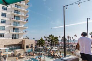 A vibrant beach scene with palm trees, a pool area, and people enjoying the sun. An aircraft leaves a trail in the clear blue sky.