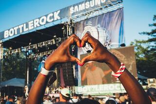 Hands form a heart shape in the foreground, while a lively concert with a large stage and audience unfolds in the background.