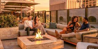 A group of friends enjoy drinks and laughter around a fire pit, illuminated by a warm sunset on a stylish rooftop terrace.