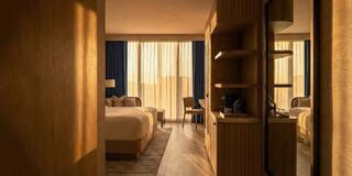 A warm, inviting hotel room features a spacious bed, elegant furnishings, and soft light filtering through sheer curtains.