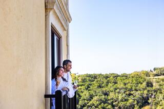 A couple enjoys drinks on a balcony, smiling and looking out over lush greenery with a clear blue sky in the background.