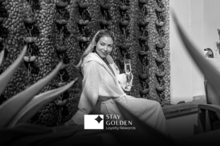 A woman in a cozy robe enjoys a drink, seated by a decorative water feature, surrounded by greenery and relaxation.