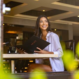 A woman smiles while holding a menu at a cozy café, enjoying a relaxed atmosphere with greenery around her.