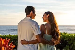 A couple stands together, gazing at the ocean sunset, surrounded by lush greenery, and radiating joy and connection.