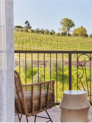 A balcony scene features a woven chair and side table, overlooking lush vineyards and a distant sculpture against a clear blue sky.