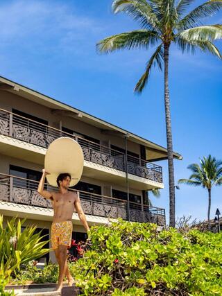 A young man with a surfboard walks through lush greenery towards a beach, surrounded by palm trees and a tropical resort backdrop.