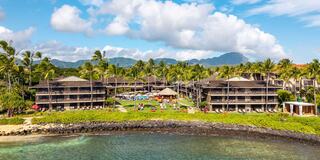A tropical resort features palm trees, multiple buildings, a pool area, and a scenic backdrop of mountains under a blue sky.