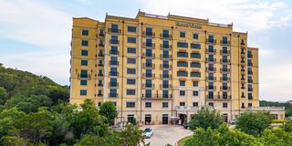 A large, elegant hotel features a golden facade and multiple balconies, set against a backdrop of lush greenery and hills.