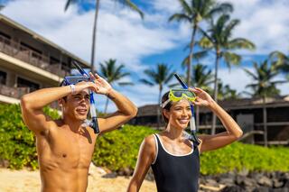 Two young adults, ready for snorkeling, smile as they stand on a sandy beach under a bright sky, with palm trees in the background.